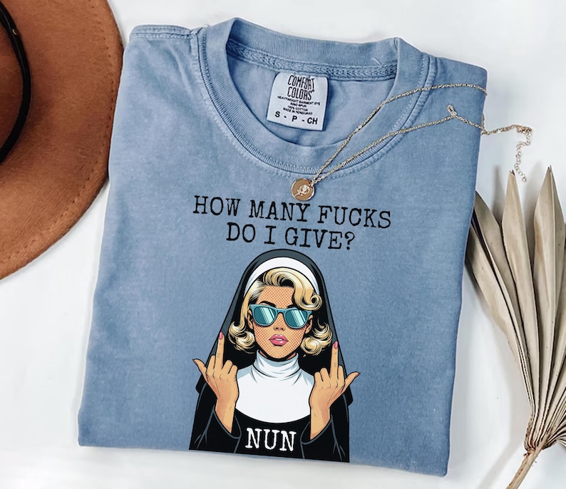 May include: A light blue t-shirt with the text "HOW MANY FUCKS DO I GIVE?" and a graphic of a nun wearing sunglasses and giving the middle finger. A gold necklace with a pendant is draped across the shirt.