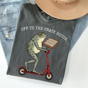 May include: Dark grey t-shirt with the text "OFF TO THE CRACK HOUSE" above a cartoon frog riding a red scooter and holding a tray of waffles. The shirt is laid on top of blue jeans and a tan hat.