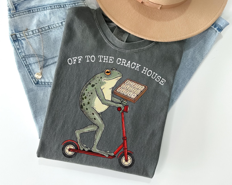 May include: Dark grey t-shirt with the text "OFF TO THE CRACK HOUSE" above a cartoon frog riding a red scooter and holding a tray of waffles. The shirt is laid on top of blue jeans and a tan hat.