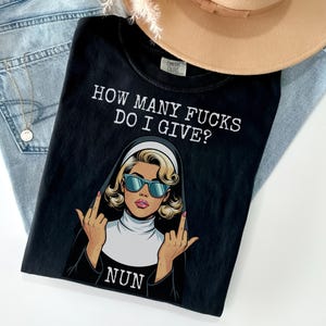 May include: Black t-shirt with the text "HOW MANY FUCKS DO I GIVE?" and a graphic of a nun wearing sunglasses and giving the middle finger. The shirt is on top of a pair of blue jeans and a tan hat.