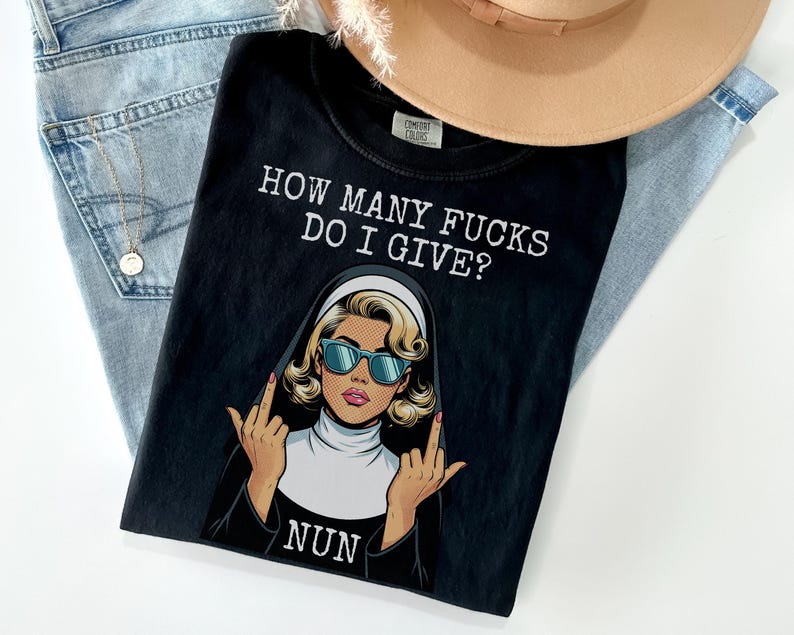 May include: Black t-shirt with the text "HOW MANY FUCKS DO I GIVE?" and a graphic of a nun wearing sunglasses and giving the middle finger. The shirt is on top of a pair of blue jeans and a tan hat.