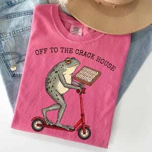 May include: A pink t-shirt with the text "OFF TO THE CRACK HOUSE" and a cartoon frog riding a red scooter while holding a tray of what appears to be a keyboard. The shirt is folded on top of blue jeans and a tan hat.