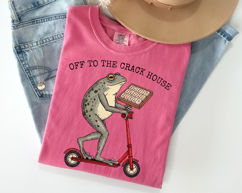 May include: A pink t-shirt with the text "OFF TO THE CRACK HOUSE" and a cartoon frog riding a red scooter while holding a tray of what appears to be a keyboard. The shirt is folded on top of blue jeans and a tan hat.