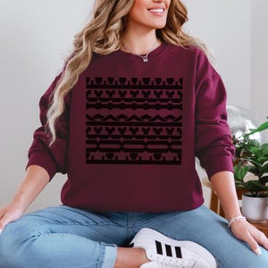 May include: Burgundy sweatshirt with a black geometric pattern. The design features repeating shapes and lines. The sweatshirt is a solid color and has a crew neck. The model is wearing blue jeans and white sneakers.