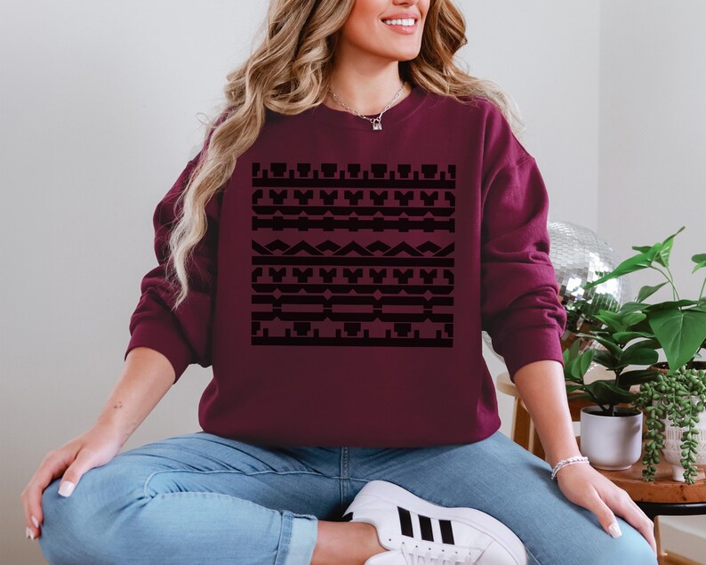 May include: Burgundy sweatshirt with a black geometric pattern. The design features repeating shapes and lines. The sweatshirt is a solid color and has a crew neck. The model is wearing blue jeans and white sneakers.