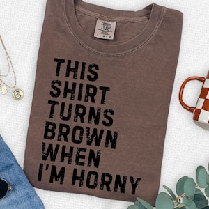May include: A brown t-shirt with the text "THIS SHIRT TURNS BROWN WHEN I'M HORNY" in black. The shirt is laid flat on a white surface, with a mug and jewelry visible in the background.