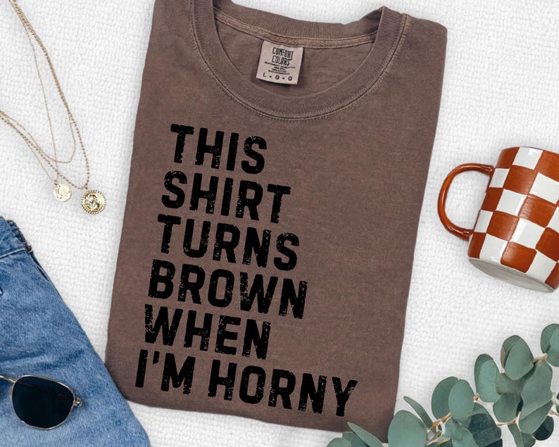 May include: A brown t-shirt with the text "THIS SHIRT TURNS BROWN WHEN I'M HORNY" in black. The shirt is laid flat on a white surface, with a mug and jewelry visible in the background.
