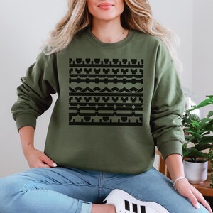 May include: Olive green sweatshirt with a black geometric pattern across the front. The long-sleeved top is made of a soft material. The design features various shapes and lines in a symmetrical arrangement.