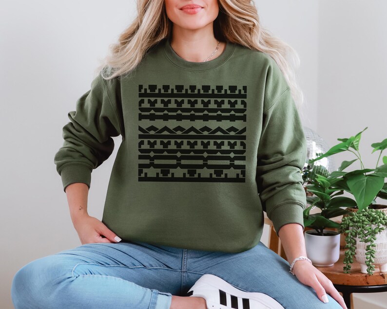 May include: Olive green sweatshirt with a black geometric pattern across the front. The long-sleeved top is made of a soft material. The design features various shapes and lines in a symmetrical arrangement.