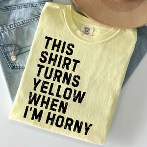 May include: A light yellow t-shirt with the text "THIS SHIRT TURNS YELLOW WHEN I'M HORNY" in black. The shirt is folded on top of a pair of blue jeans and a tan hat.