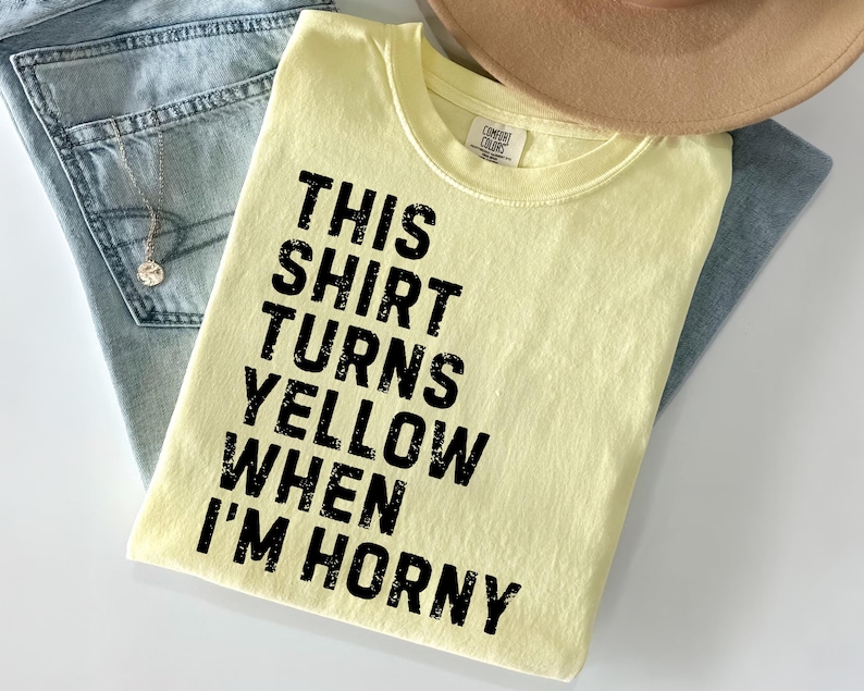 May include: A light yellow t-shirt with the text "THIS SHIRT TURNS YELLOW WHEN I'M HORNY" in black. The shirt is folded on top of a pair of blue jeans and a tan hat.