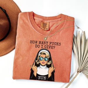May include: Coral-colored t-shirt with the phrase "HOW MANY FUCKS DO I GIVE?" above a cartoon nun wearing sunglasses and giving the middle finger. A gold necklace and a brown hat are also in the image.