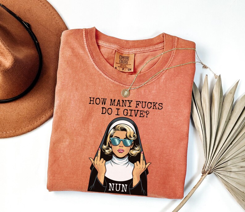 May include: Coral-colored t-shirt with the phrase "HOW MANY FUCKS DO I GIVE?" above a cartoon nun wearing sunglasses and giving the middle finger. A gold necklace and a brown hat are also in the image.