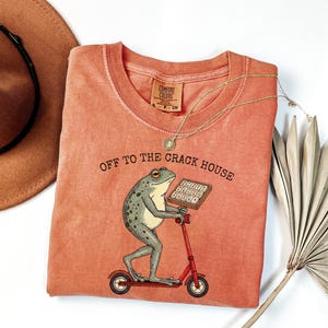 May include: Coral-colored t-shirt with a frog graphic riding a red scooter and holding a calculator. The text "OFF TO THE CRACK HOUSE" is printed above the frog. A gold necklace and a brown hat are also in the image.