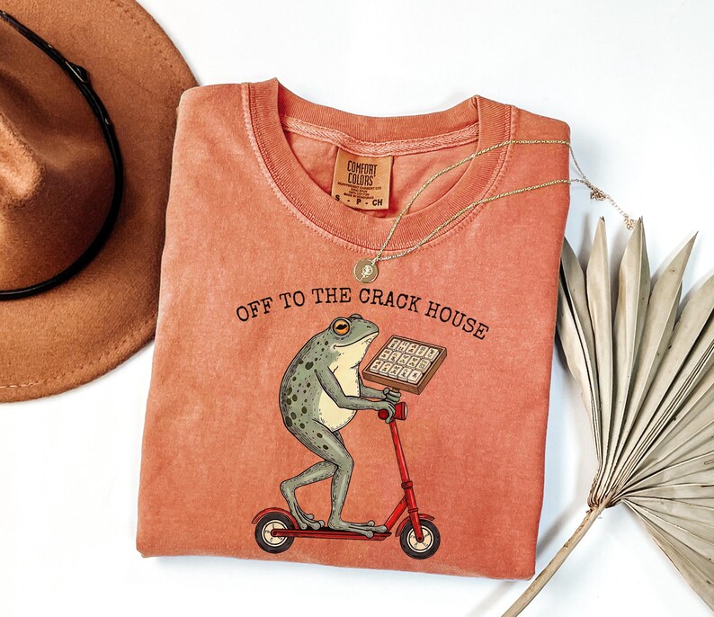 May include: Coral-colored t-shirt with a frog graphic riding a red scooter and holding a calculator. The text "OFF TO THE CRACK HOUSE" is printed above the frog. A gold necklace and a brown hat are also in the image.