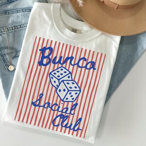 May include: A white t-shirt featuring a red vertical striped rectangular graphic with two blue dice in the center. The text "Bunco Social Club" is printed in blue script font over the stripes. The shirt has a crew neckline and short sleeves.