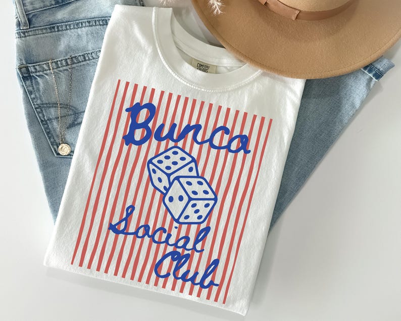 May include: A white t-shirt featuring a red vertical striped rectangular graphic with two blue dice in the center. The text "Bunco Social Club" is printed in blue script font over the stripes. The shirt has a crew neckline and short sleeves.
