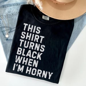 May include: Black t-shirt with the text "THIS SHIRT TURNS BLACK WHEN I'M HORNY" in white. The shirt is laid on top of a pair of light blue jeans and a tan hat.