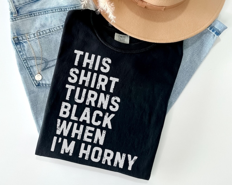 May include: Black t-shirt with the text "THIS SHIRT TURNS BLACK WHEN I'M HORNY" in white. The shirt is laid on top of a pair of light blue jeans and a tan hat.