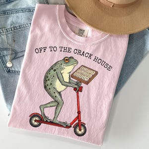 May include: Pink t-shirt with the text "OFF TO THE CRACK HOUSE" and a cartoon frog riding a red scooter while holding a tray of waffles. The shirt is laid on top of blue jeans and a tan hat.