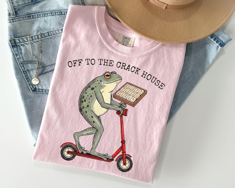 May include: Pink t-shirt with the text "OFF TO THE CRACK HOUSE" and a cartoon frog riding a red scooter while holding a tray of waffles. The shirt is laid on top of blue jeans and a tan hat.
