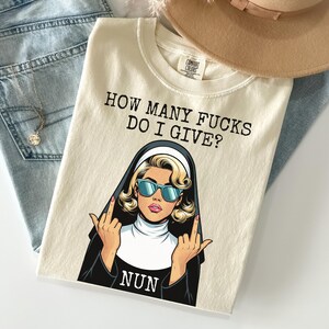 May include: Cream-colored t-shirt with the text "HOW MANY FUCKS DO I GIVE?" and a graphic of a nun wearing sunglasses and giving the middle finger. A tan hat and blue jeans are in the background.