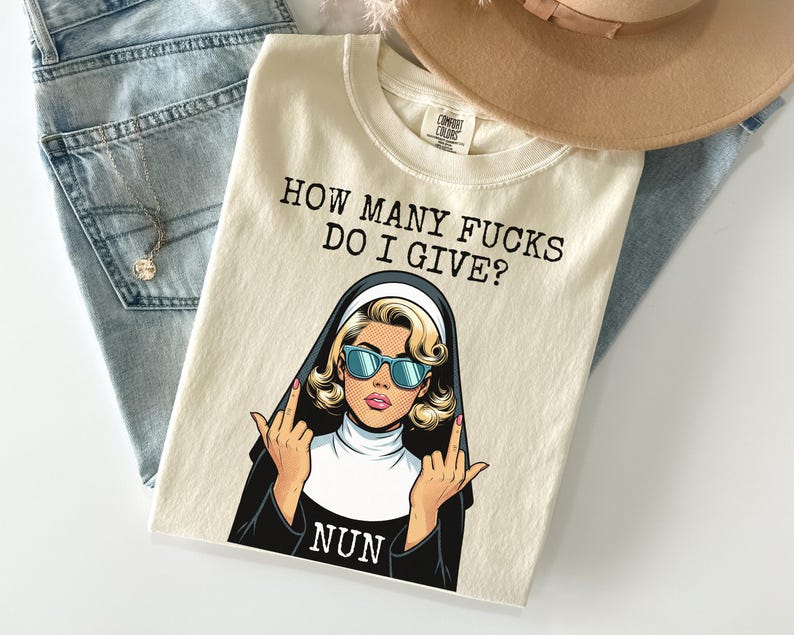 May include: Cream-colored t-shirt with the text "HOW MANY FUCKS DO I GIVE?" and a graphic of a nun wearing sunglasses and giving the middle finger. A tan hat and blue jeans are in the background.