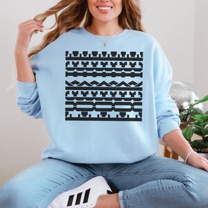 May include: A light blue sweatshirt with a black geometric pattern. The pattern consists of various shapes, including rectangles, triangles, and other abstract designs. The sweatshirt is long-sleeved and has a crew neck.