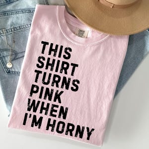 May include: A light pink t-shirt with the text "THIS SHIRT TURNS PINK WHEN I'M HORNY" in black. The shirt is laid on top of a pair of light blue jeans and a tan hat.