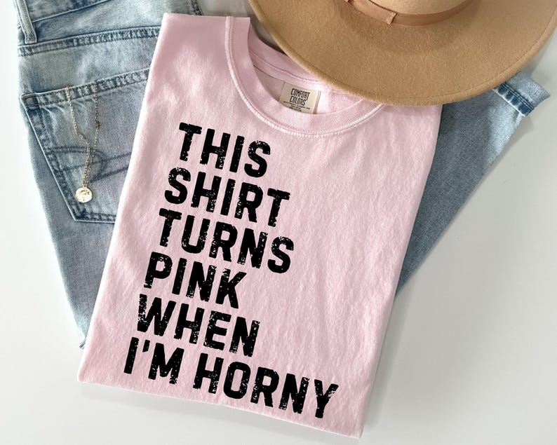 May include: A light pink t-shirt with the text "THIS SHIRT TURNS PINK WHEN I'M HORNY" in black. The shirt is laid on top of a pair of light blue jeans and a tan hat.