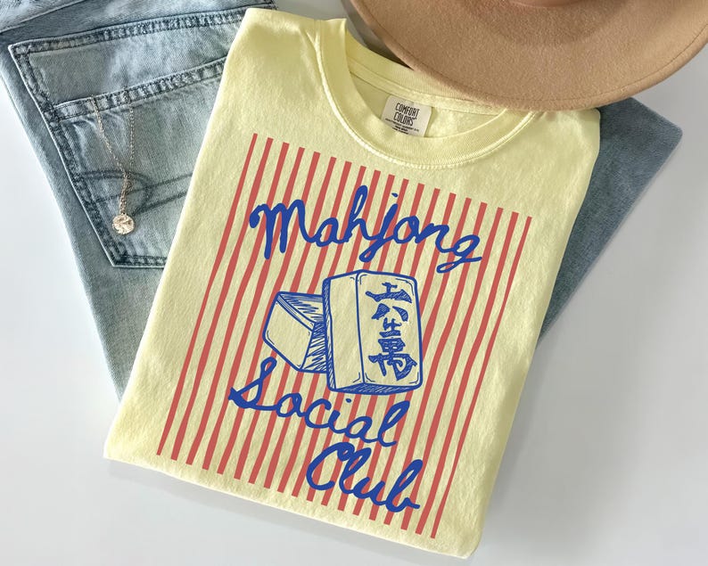 May include: A light yellow t-shirt with a red and white striped design and the words "Mahjong Social Club" in blue. The shirt is laid on top of a pair of light blue jeans and a tan hat.