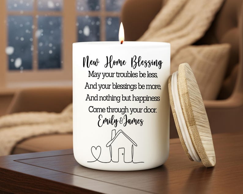 Puede incluir: Una vela blanca con tapa de madera, con la inscripci&oacute;n "New Home Blessing" y una ilustraci&oacute;n de una casa. La vela incluye los nombres "Emily & James" y un s&iacute;mbolo de coraz&oacute;n. La vela est&aacute; encendida, con una llama visible.