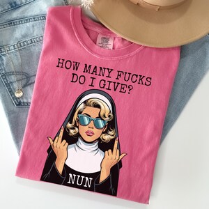 May include: Pink t-shirt with the text "HOW MANY FUCKS DO I GIVE?" and a graphic of a nun wearing sunglasses and giving the middle finger. The shirt is folded on top of blue jeans and a tan hat.