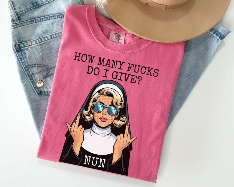 May include: Pink t-shirt with the text "HOW MANY FUCKS DO I GIVE?" and a graphic of a nun wearing sunglasses and giving the middle finger. The shirt is folded on top of blue jeans and a tan hat.