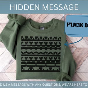 May include: An olive green sweatshirt with a black geometric pattern across the chest. A blue box with the text "FUCK ICE" is in the upper right corner. The text "HIDDEN MESSAGE" is at the top, and "SEND US A MESSAGE WITH ANY QUESTIONS, WE ARE HERE TO HELP!" is at the bottom.