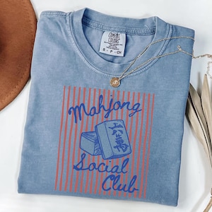May include: A light blue t-shirt with the words "Mahjong Social Club" in a red and blue design. The shirt has a crew neck and short sleeves. A gold necklace with a pendant is on the shirt.