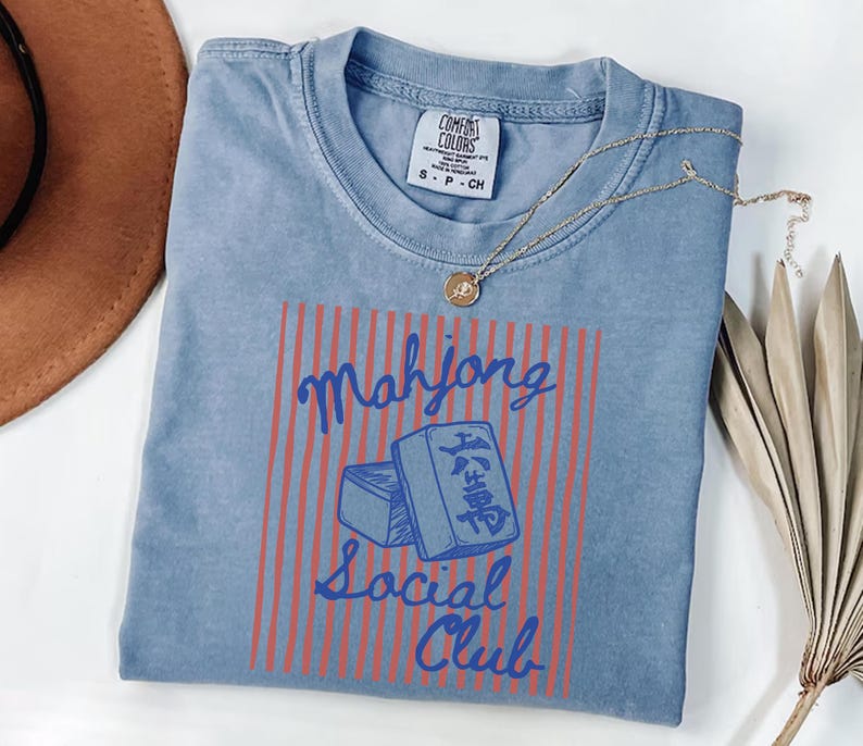 May include: A light blue t-shirt with the words "Mahjong Social Club" in a red and blue design. The shirt has a crew neck and short sleeves. A gold necklace with a pendant is on the shirt.
