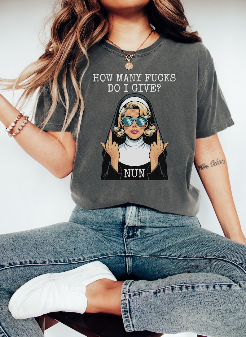 May include: Dark gray t-shirt with the text "HOW MANY FUCKS DO I GIVE?" above an illustration of a nun wearing sunglasses and giving the middle finger. The word "NUN" is below the illustration.