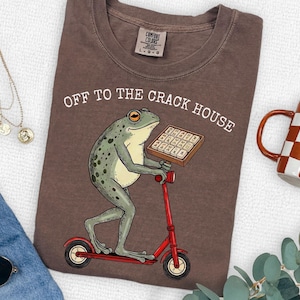 May include: Brown t-shirt with the text "OFF TO THE CRACK HOUSE". The shirt features a cartoon frog riding a red scooter and holding a tray of what appears to be chocolate bars. A checkered mug is visible in the background.