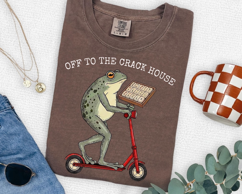 May include: Brown t-shirt with the text "OFF TO THE CRACK HOUSE". The shirt features a cartoon frog riding a red scooter and holding a tray of what appears to be chocolate bars. A checkered mug is visible in the background.
