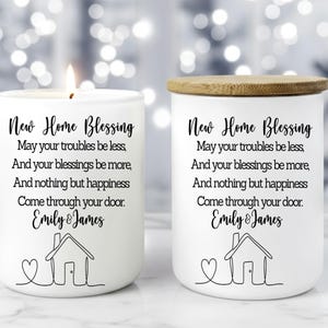 Puede incluir: Dos velas de cer&aacute;mica blanca con tapas de madera. Una vela est&aacute; encendida. Cada vela tiene el texto "New Home Blessing" y un mensaje personalizado con una ilustraci&oacute;n de una casa. Un regalo de inauguraci&oacute;n.