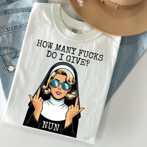 May include: White t-shirt with the text "HOW MANY FUCKS DO I GIVE?" above a pop art style illustration of a nun wearing sunglasses and giving the middle finger. The word "NUN" is below the illustration.