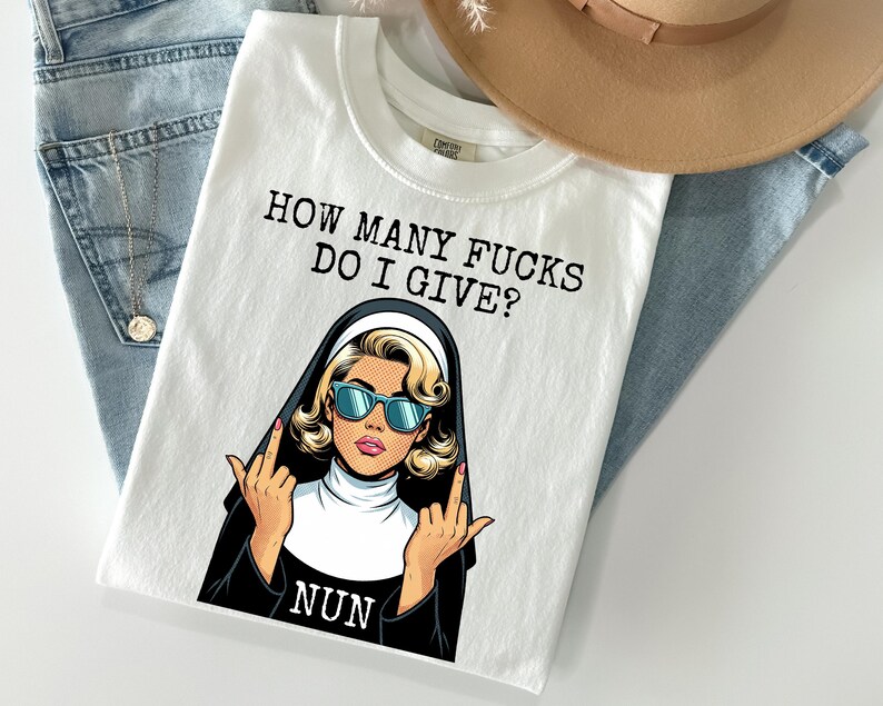 May include: White t-shirt with the text "HOW MANY FUCKS DO I GIVE?" above a pop art style illustration of a nun wearing sunglasses and giving the middle finger. The word "NUN" is below the illustration.