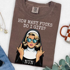 May include: A brown t-shirt featuring a graphic of a nun wearing blue sunglasses and giving the middle finger with both hands. The text above reads HOW MANY FUCKS DO I GIVE? and the word NUN is printed below the graphic in white lettering.