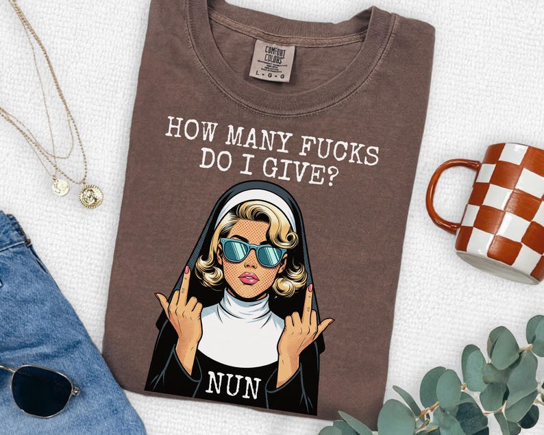 May include: A brown t-shirt featuring a graphic of a nun wearing blue sunglasses and giving the middle finger with both hands. The text above reads HOW MANY FUCKS DO I GIVE? and the word NUN is printed below the graphic in white lettering.