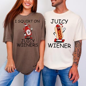 May include: Two t-shirts with humorous graphics. One is brown with a ketchup bottle character and the text "I SQUIRT ON JUICY WIENERS." The other is cream-colored with a hot dog character and the text "JUICY WIENER."