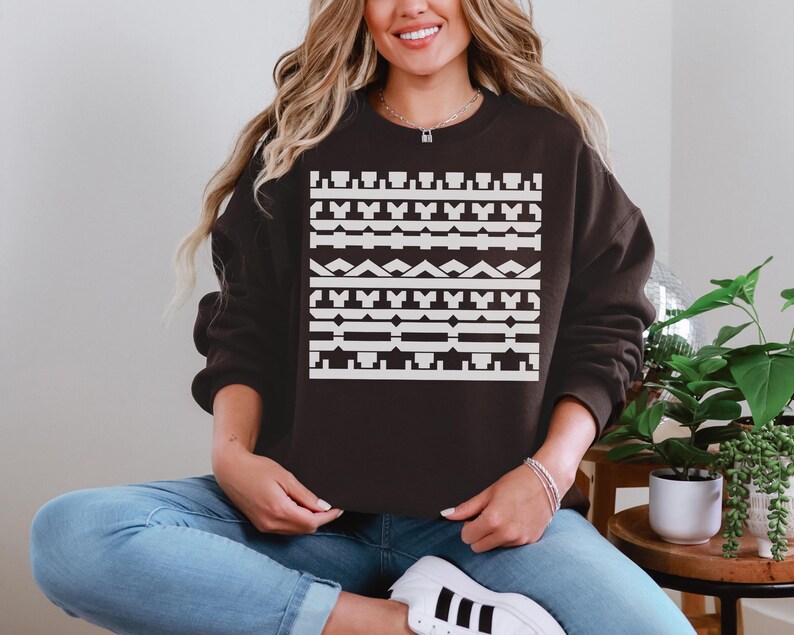May include: A dark brown sweatshirt with a geometric white pattern across the front. The pattern consists of various shapes, including rectangles, triangles, and lines. The sweatshirt is a casual style, suitable for everyday wear.