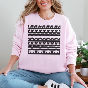 May include: A light pink sweatshirt featuring a black geometric pattern across the chest. The pattern consists of various shapes and lines arranged in horizontal rows. The sweatshirt is a solid light pink color.