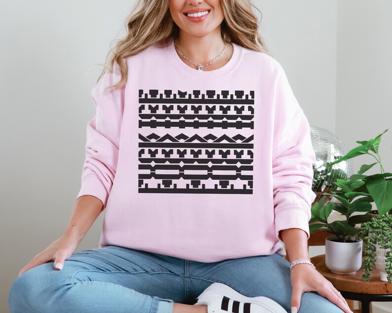 May include: A light pink sweatshirt featuring a black geometric pattern across the chest. The pattern consists of various shapes and lines arranged in horizontal rows. The sweatshirt is a solid light pink color.