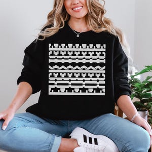 May include: Black sweatshirt with a white geometric pattern across the chest. The pattern consists of various shapes and lines. The model is wearing blue jeans and white sneakers. The sweatshirt is a casual style.